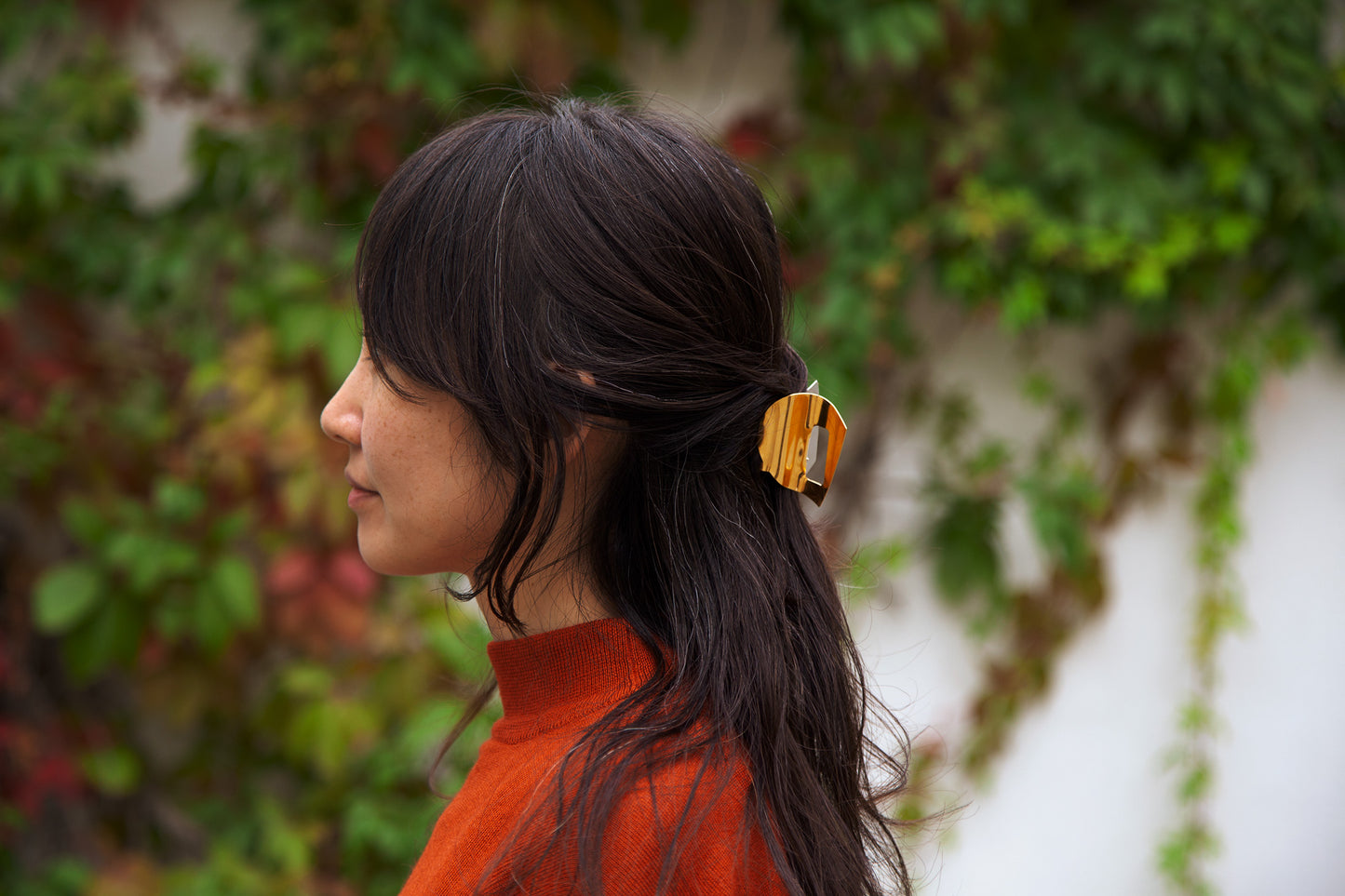 Woman with long dark hair tied back with gold-plated claw clip MIRA by CLINQ wearing an orange top, standing against a blurred natural background.